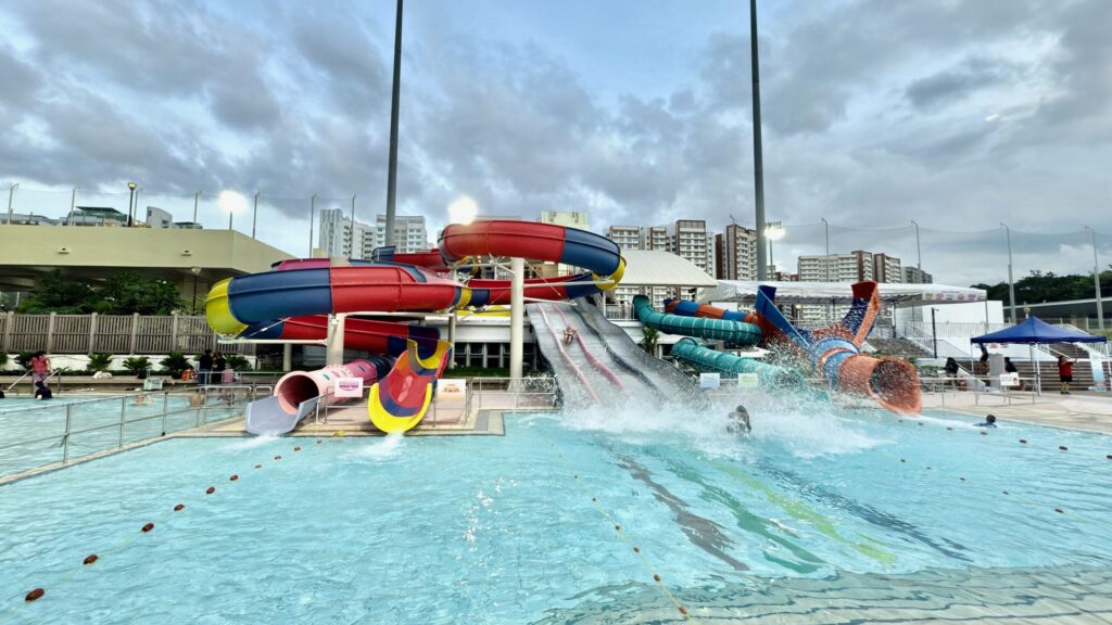 Image 2: Multiple colourful water slides at Sengkang Swimming Complex splashing into a feature pool with swimmers in action.